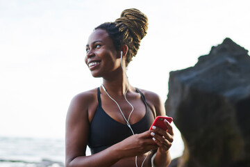 Cheerful afro american lover of sport looking away while installing music app on phone for morning workout on coasline of tropical island.Positive dark skinned female with earphones preparing for run