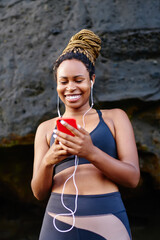 Cheerful dark skinned female jogger happy from cool music on radio for morning workout on coastline.Positive young woman listening audio from player in headphones connected to cellular before training