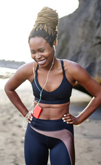 Cheerful dark skinned young woman dressed in stylish sport wear laughing during morning workout and listening funny audio songs in eraphones.Afro american female training on seashore enjoying music