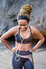 Cheerful afro american young woman dressed in sport clothes laughing during workout on coastline.Positive dark skinned female in active wear listening audio songs in earphones connected to smartphone
