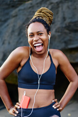 Portrait of emotional positive sportive woman dressed in active clothes enjoying funny music from player on smartphone.Cheerful afro american female listening songs in earphones before sport training