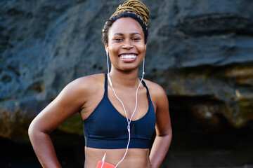 happy afro american young woman smiling at camera while litening audio songs from music application in earphones.Cheerful dark skinned jogger dressed in sport top smiling at camera
