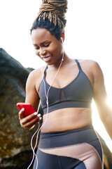 Cheerful afro american sportive woman in earphones connected to smartphone choosing music application for download on smartphone and morning training.Positive female in active wear and phone in hands