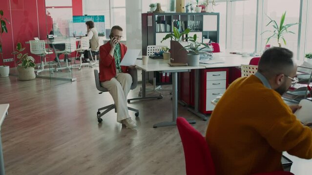 Full zoom-in shot of Caucasian employees working in office of creative advertising agency - man sitting at desk studying financial report, woman in red jacket talking with client on mobile phone