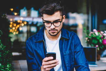 Pensive young man in eyewear reading notification on smartphone while updating software,...