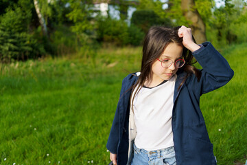 A little girl in glasses, a T-shirt and a jacket scratches her head while standing on a spring...