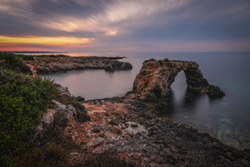 Rocky coastline with a natural arch at punta Asparano, near Siracusa. Sunrise time, long exposure picture. June 2023