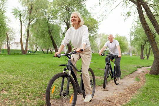 elderly senior couple rides bicycle in the park in the summer and smiles, old gray-haired man and woman are actively resting outdoors, old people practice cycling in forest