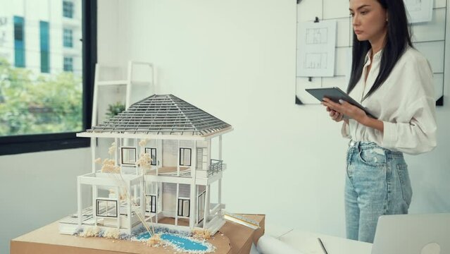 Beautiful caucasian architect focus on using tablet to check house model on meeting table with blueprint, house model and architectural document scatter around at meeting room. Immaculate.