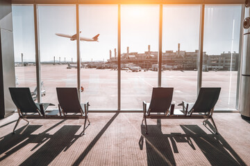 View from an airport terminal window showing chairs facing the runway. A plane is taking off...