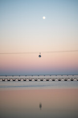 A vertical scenic view of a cable car traveling over a calm body of water with a bridge in the background, captured during a serene sunset. The sky transitions from pink to blue with the moon visible