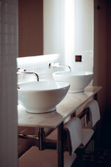 Close-up vertical shot of a modern bathroom with two white ceramic vessel sinks and chrome faucets on a marble countertop. The background features white tiled walls and neatly folded towels