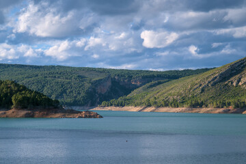 The image shows a serene lake surrounded by green hills and forested mountains. 