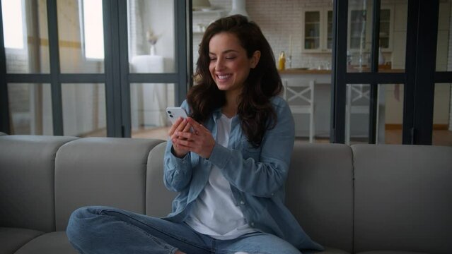Happy smiling excited rejoice joyful winner girl Caucasian female woman user influencer win achieve success good news mobile phone online app smartphone hugging excitement relaxing at home couch sofa