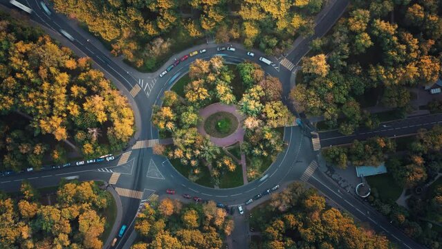 Top down time-lapse aerial view of a park with a roundabout intersection with slow camera rotation, beautiful sunset illuminates trees. Cars move fast along the road during an autumn traffic jam.