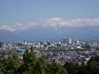 Obraz premium 5月新緑の木々と富山市街地の後方に見える残雪の北アルプス薬師岳