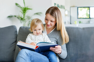 Young mother at home, reading a book with her baby daughter. Early learning and motherhood concept.