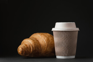 Paper cup of hot coffee with paper lid next to freshly baked croissant on the black background. Mock up with copy space for a free text