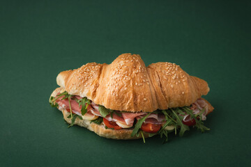 Freshly baked croissant with iberian ham, tomatoes and salad filling on dark green background. Mock...