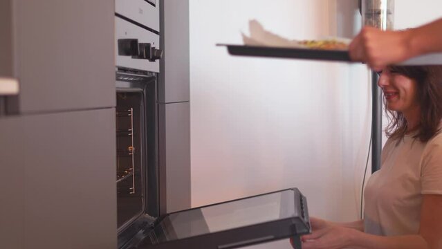 Woman opening oven while friend placing tray with salmon to be grilled in it at home. Female friends have gathered for lunch at home. 