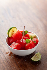 A white ceramic bowl filled decoratively with Organic watermelon (citrullus Lanatus) cubes, lemon wedge, and mint leaves, on a wooden background.