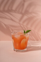 Summer refreshing drink with iced peach juice or fruit alcohol cocktail in glass on vibrant peachy pink background with tropical floral sunlight shadows