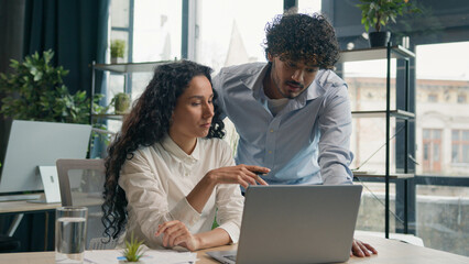 Indian Arabian male coach mentor leader businessman help Hispanic businesswoman assistance new employee teaching intern explaining online software discuss marketing data on laptop business teamwork