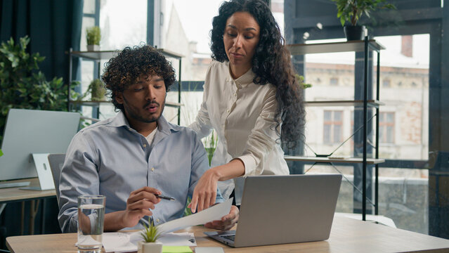 Arabian female leader coach help intern explain paperwork and financial data business strategy consult client. Smiling Indian colleague businessman talk with businesswoman corporate teamwork at office