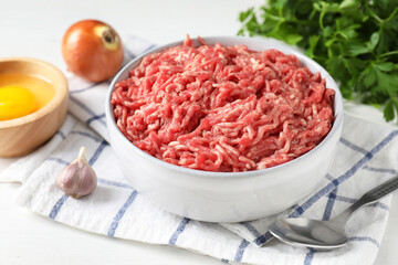 Raw ground meat in bowl, garlic, egg, parsley and spoon on white table, closeup