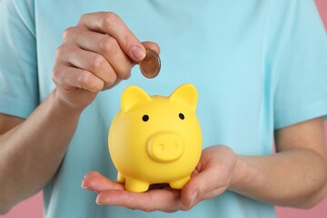 Woman putting coin into yellow piggy bank on pink background, closeup