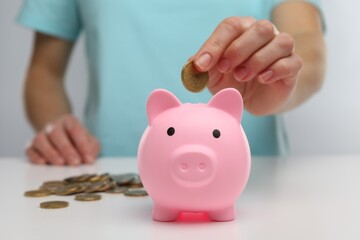 Woman putting coin into pink piggy bank at white table, closeup