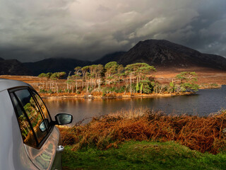 Side of silver car parked off road, stunning scenery of Connemara with lake, island, mountains and dramatic sky in the background. County Galway, Ireland. Travel and rent car concept.
