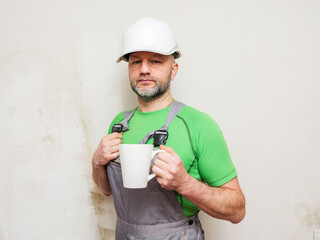 A man in a green shirt and gray overalls holding a cup of tea or coffee. Male construction worker with grey beard in white safety plastic helmet. Light color background.