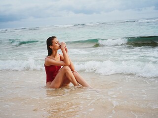 A young woman in a red swimsuit sits on the sand with a beautiful sun tan and looks out at the ocean in the tropics on an island beach