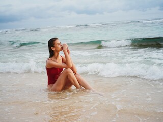 A young woman in a red swimsuit sits on the sand with a beautiful sun tan and looks out at the...