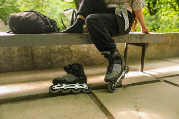 A teenage girl on roller skates sits on a bench in a city park and adjusts her equipment, her...