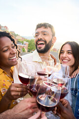 Vertical. Cheerful international friends toasting glasses of red wine together laughing at terrace bar outdoors. Close up cheers of smiling diverse people having fun at sunset party. Summer vibes