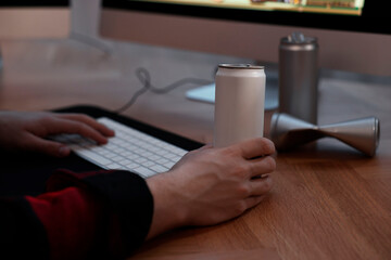 Young man with energy drink playing video game at wooden desk indoors, closeup