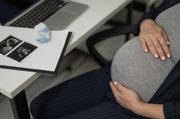 A pregnant woman works on a laptop in the office and looks at a photo from an ultrasound scan of...