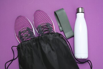 Black drawstring bag, sneakers, thermo bottle and fitness elastic band on purple background, flat lay