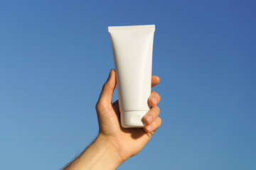 A man's hand holds a white mockup tube of cream on a blue sky background. Concept of natural...