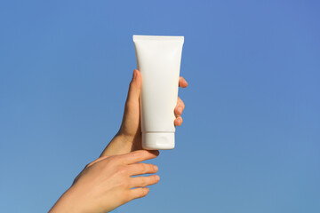 Female fragile hands hold a white mockup tube of face or body cream on a blue sky background. Skin care, beauty and spf products concept.