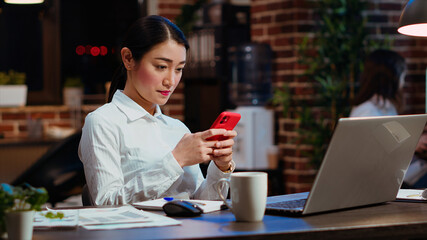 Smiling employee at work texting friends on cellphone, taking break from solving project tasks. Relaxed businesswoman using mobile phone to unwind in office late at night