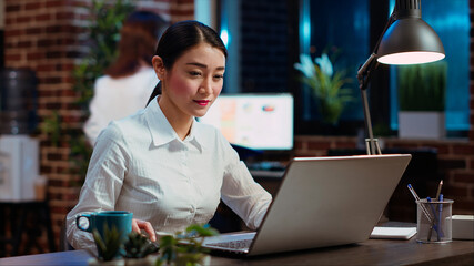 Businesswoman drinking cup of coffee while doing tasks in office overnight for company project. Employee enjoying hot beverage while looking over figures on laptop late at night, typing on keyboard
