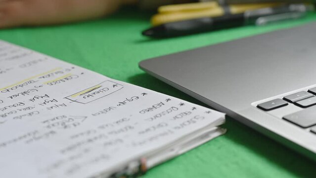 Young college student uses a pen to mark a word on a notebook next to a computer while studying in her dorm room