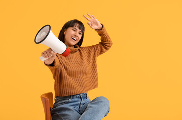 Young woman with megaphone showing OK while sitting on chair against yellow background
