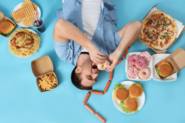 Young man with unhealthy food lying on blue background, top view. Overeating concept