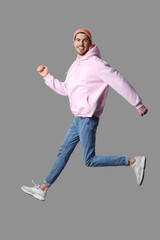 Handsome smiling young man in stylish pink hoodie and peach hat jumping on grey background