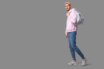 Fototapeta premium Handsome young man in stylish pink hoodie and peach hat with backpack posing on grey background