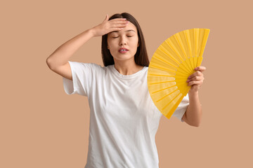 Young woman with hand fan suffering from heat on beige background